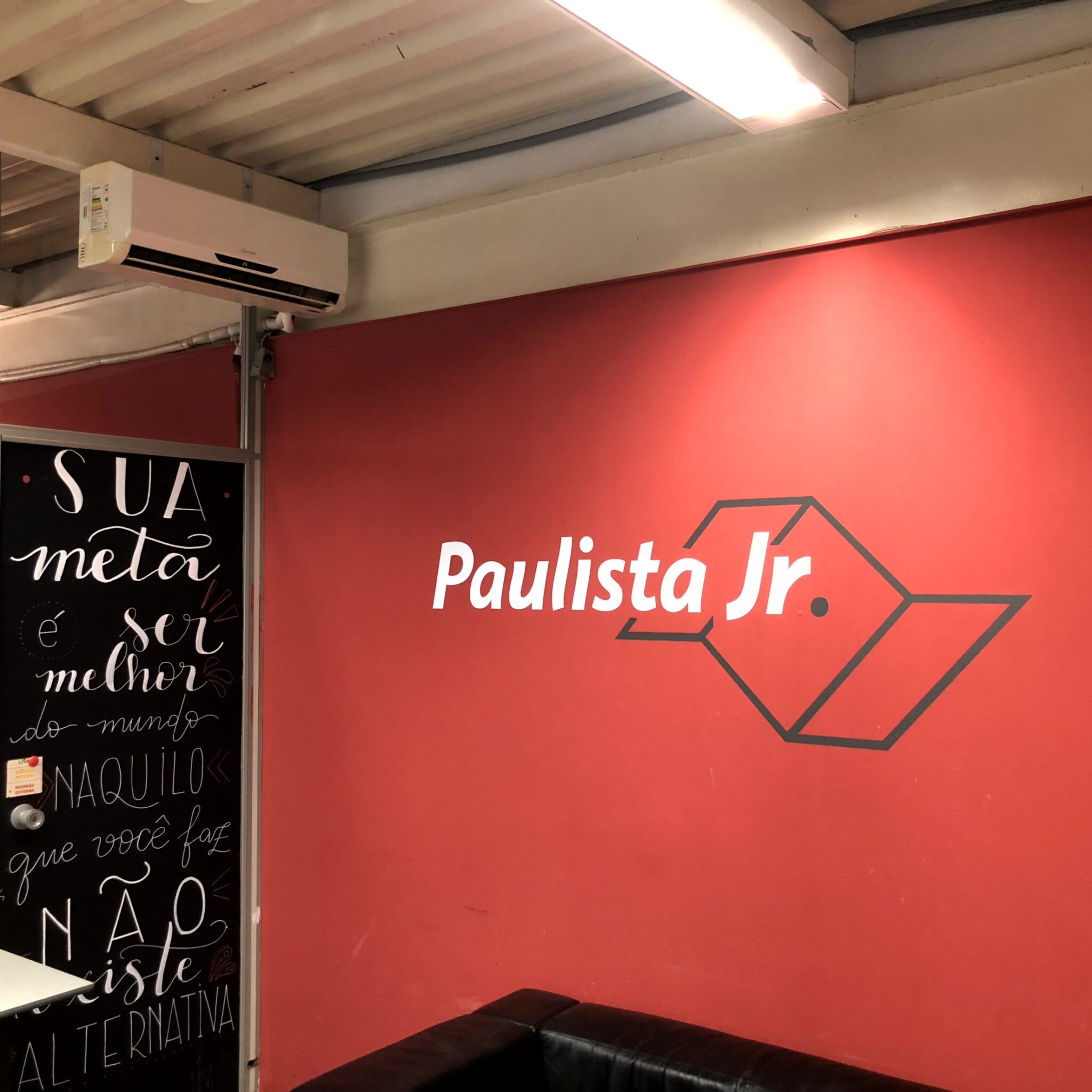 Parede vermelha com a logo da Paulista Jr, o nome da empresa escrito em branco com o simbolo do estado de São Paulo