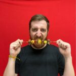 Homem branco, cabelo loiro curto, barba e bigode completo loiro, camiseta preta, segurando com ambas as mãos um palito de churrasco com pedaços de carne e legumes em frente a boca enquanto faz pose de quem vai morde-lo