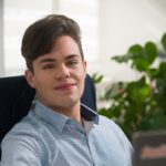 Homem branco loiro, olhos claros, cabelo penteado em topete, camisa social azul